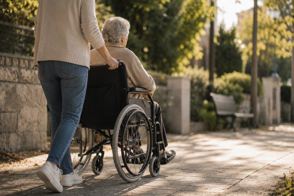 Una cuidadora passeja una dona gran en cadira de rodes per un carrer arbrat